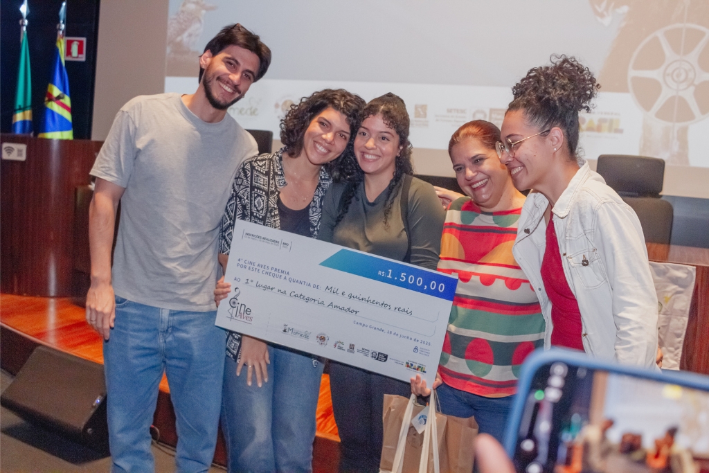 Julice, ao centro, veste blusa preta de manga longa e segura o cheque simbólico da premiação recebida no Cine Aves.. Foto: Tero Queiroz